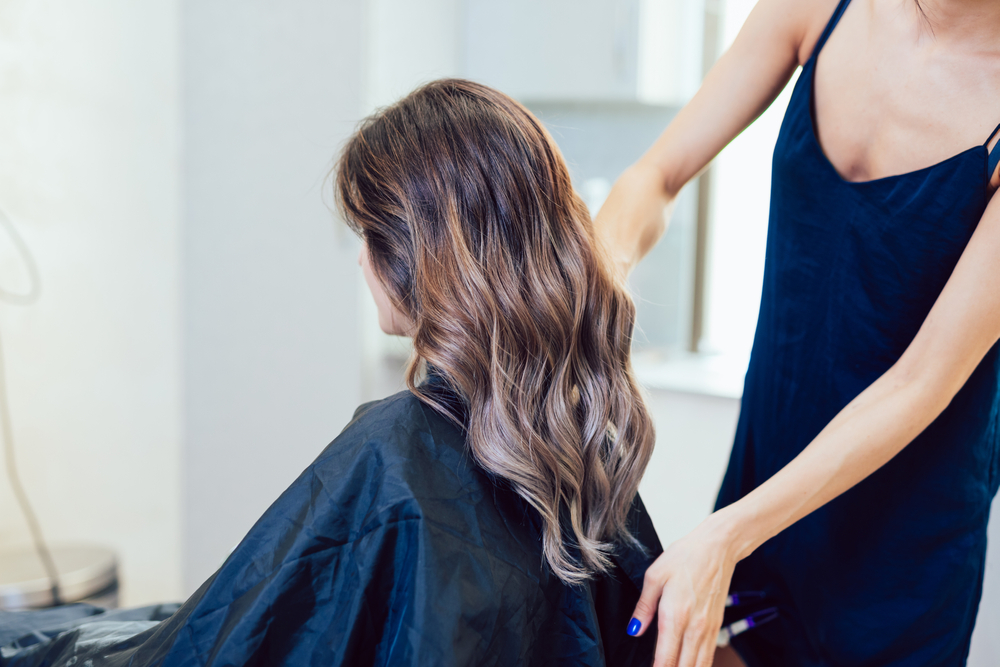 Stylist lifts finished long, curly grey ombre hair on her client sitting in a chair facing to the side with black cape over her shoulders