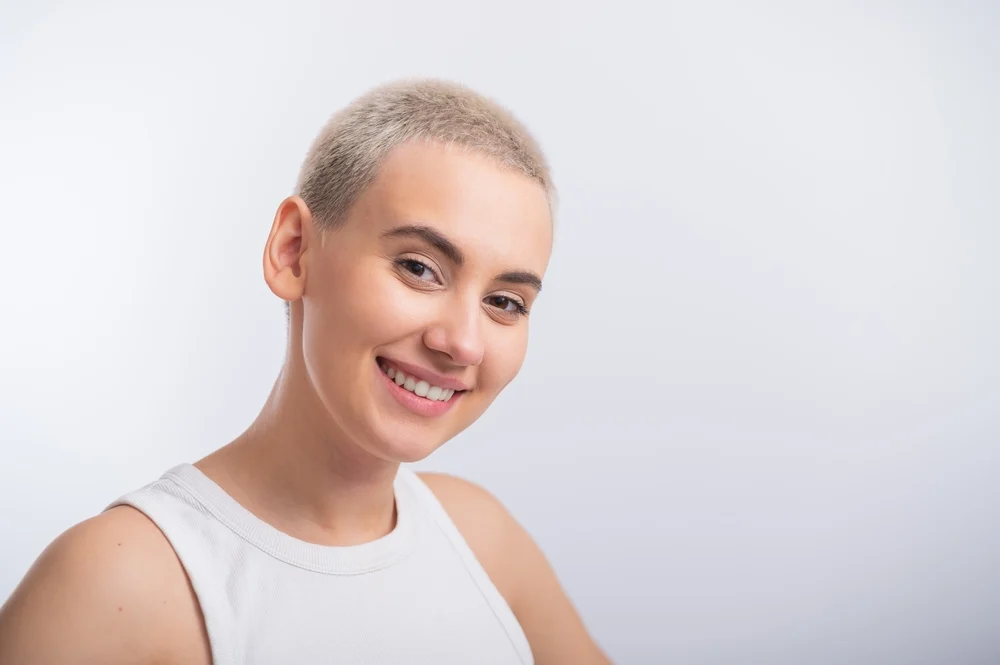 Young blonde woman in a white tank top smiles and shows off her short cropped buzz cut that perfectly suits oval face shapes in front of a gray wall