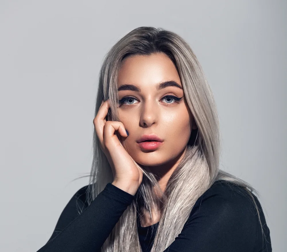 Woman looks serious as she poses with her hand against her cheek in a long-sleeved back shirt in front of a grey background to indicate what grey ombre hair is