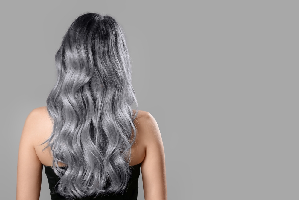 Rear view of woman standing in front of a grey wall to show an example of grey and black ombre hair color styled in waves