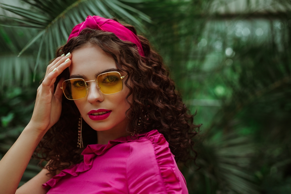 An example of cute hairstyles for curly hair is loose curls with a bright pink statement headband, shown here on a brunette model outside with greenery behind her