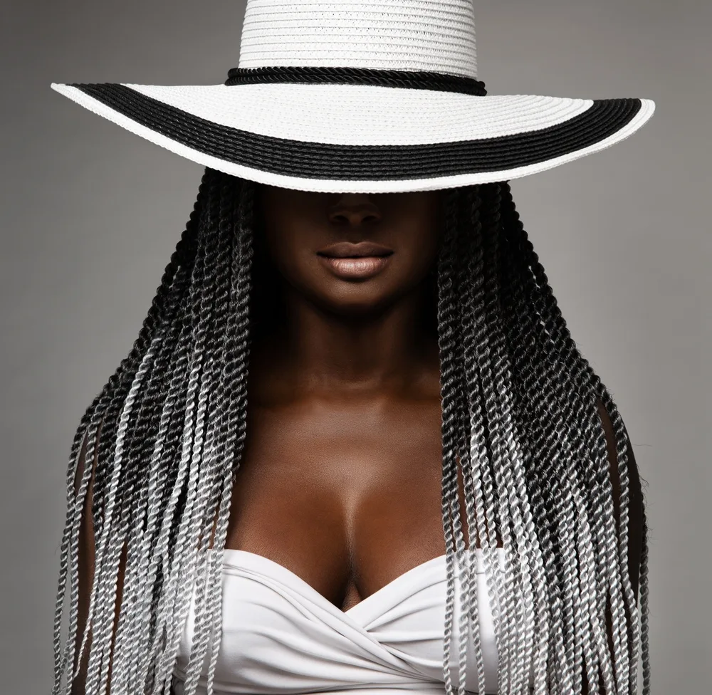African American woman wearing a big hat and white swimsuit peeks out with her grey ombre hair color showing off small twists