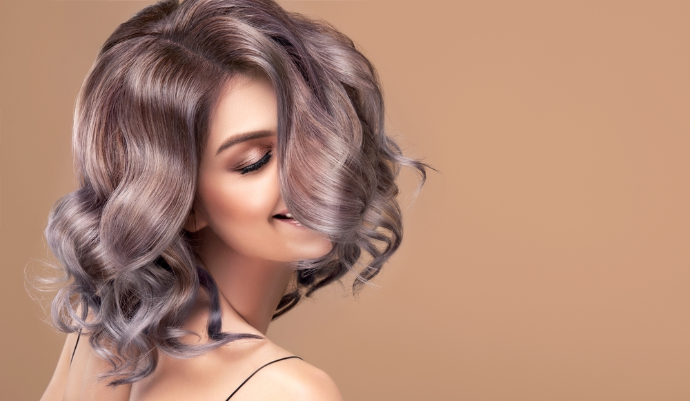Smiling woman twirls her short curly hair over her shoulder to show off grey ombre color with dark blonde at the roots