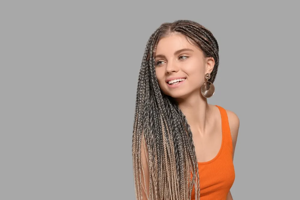 Smiling young woman in an orange tank top shows off braids over her shoulder in an ombre style from grey to blonde