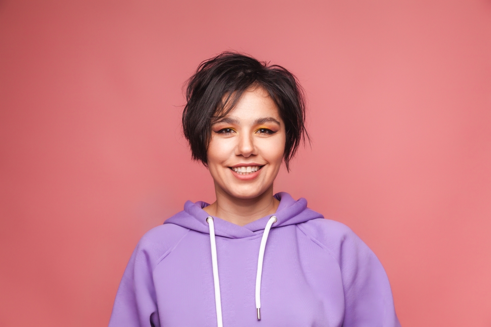 Dark-haired woman in a purple hoodie smiles with her shaggy pixie cut parted to the side, indicating one of the top short haircuts for oval faces