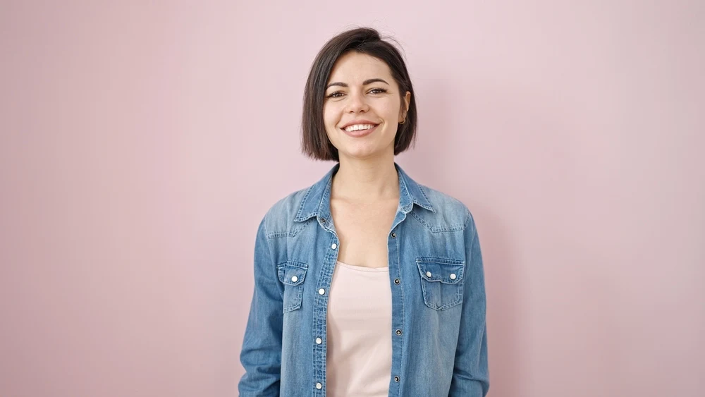 Brunette woman stands smiling in front of a pink wall with denim jacket and a short bob haircut for oval faces