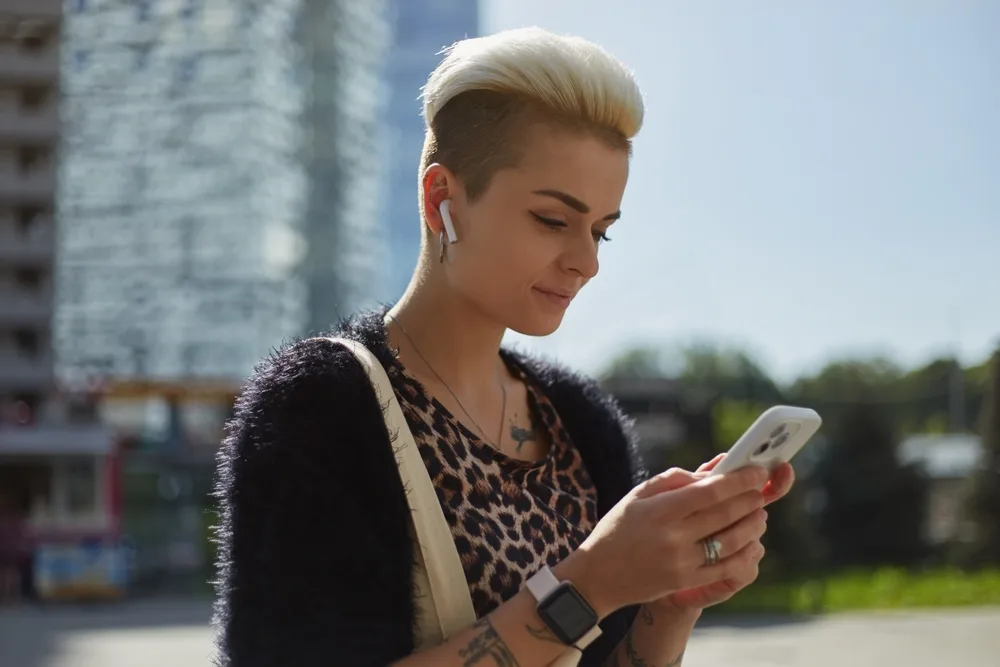 Blonde woman wears a short haircut for oval faces, a pompadour pixie undercut, while looking down at her smartphone outside