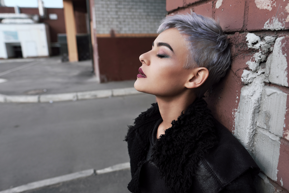 Young woman with a short pixie haircut leans back against a wall outside with a lilac silver ombre color and a black leather jacket with fluffy collar