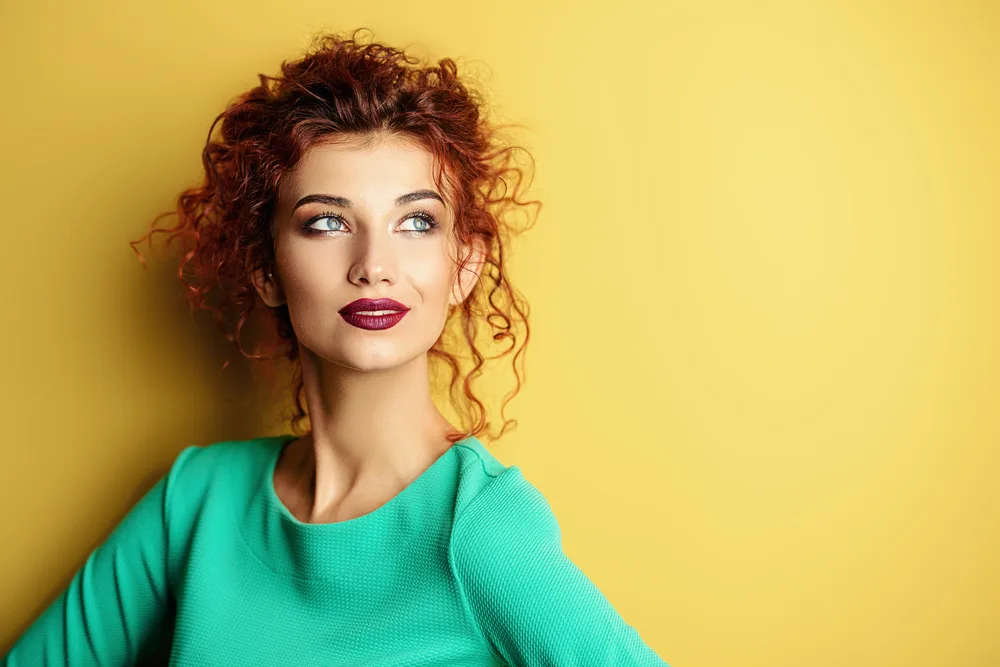 Red-haired woman with curly hair wears a loose, artsy ponytail and a teal long-sleeved top in front of a yellow background as an example of cute hairstyles for curly hair