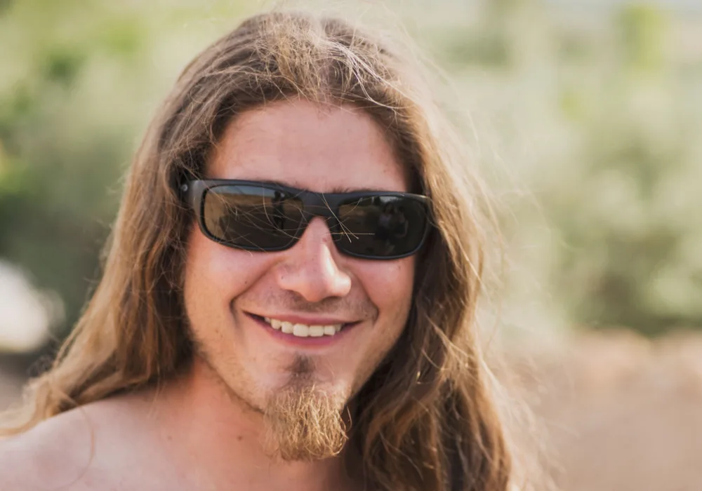 Long-haired blonde man poses outside with sunglasses on showing his long Norse skipper goatee that extends beyond the chin