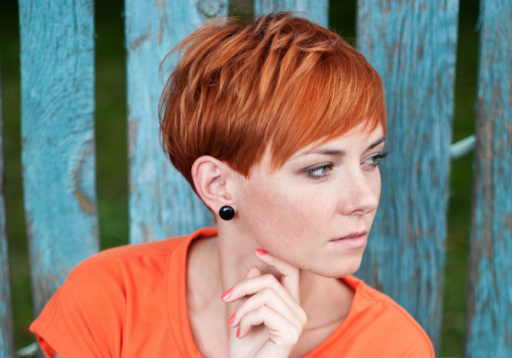 Serious woman with bright copper hair and a short pixie cut glances away in front of a wooden fence to show an example of fine hair thin hair low maintenance short hairstyles 