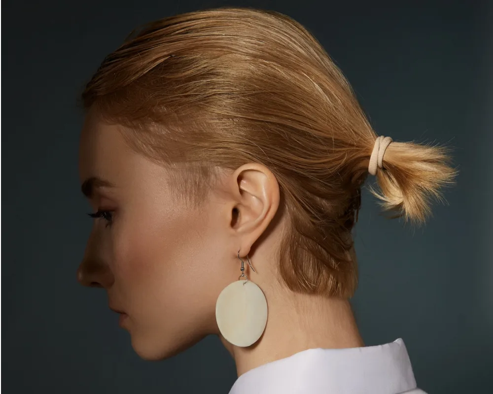Side view of blonde woman in white collared shirt showing her short half-tucked ponytail and round dangling earrings