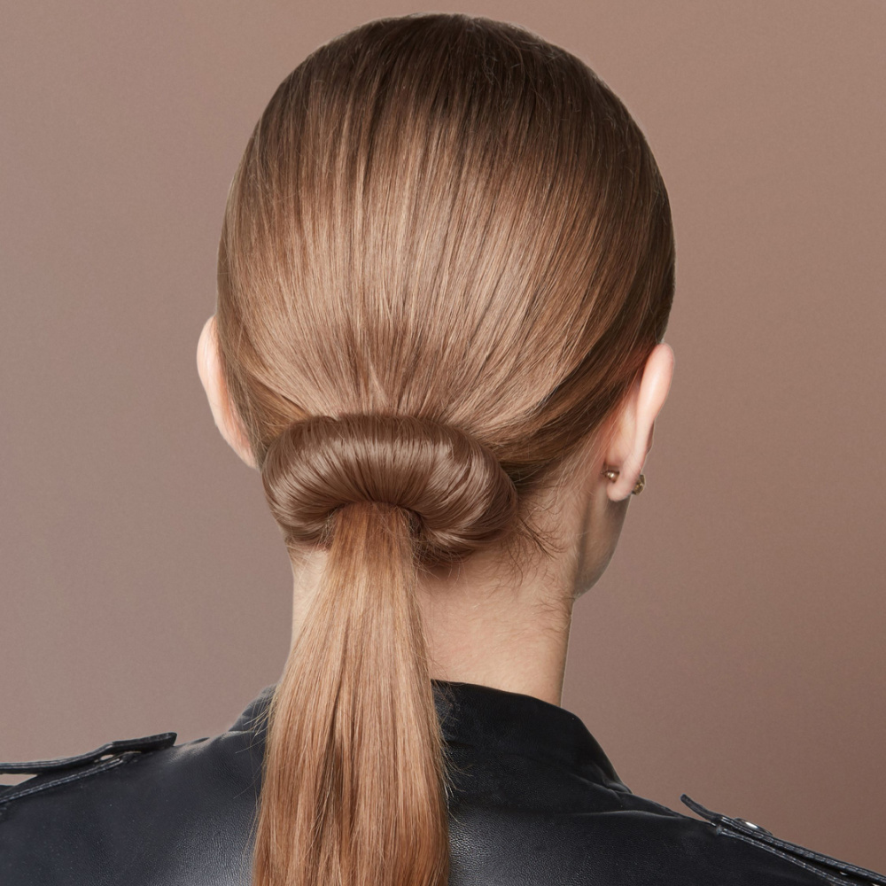 Back close-up view of a woman's ponytail hairstyle featuring a bun wrapped around loose hair in front of a light brown background