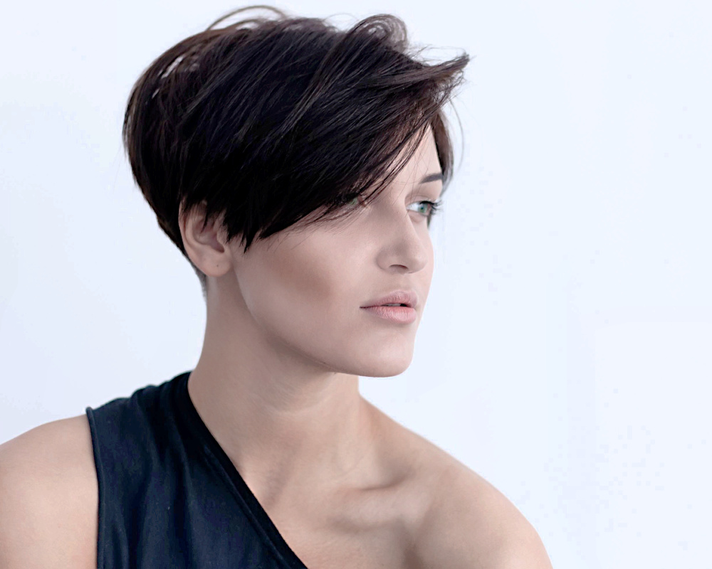 Example of fine hair thin hair low maintenance short hairstyles includes a short layered pixie cut with a side part and lots of volume, as shown on a brunette model facing away with a one-shoulder shirt