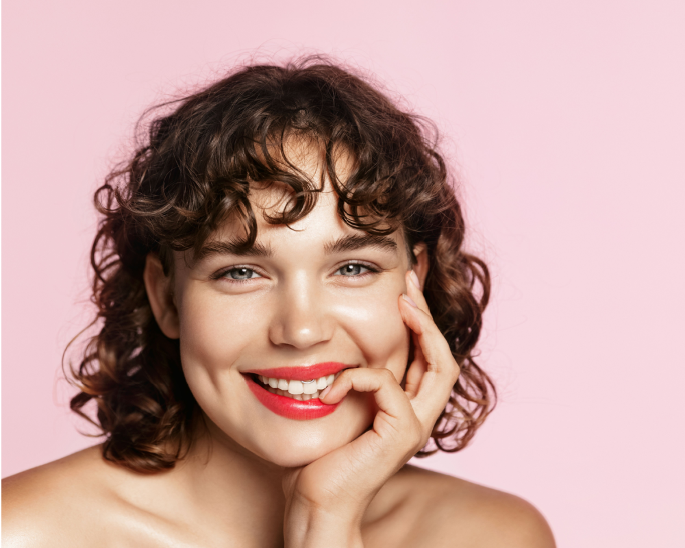 Woman with red lipstick smiles and bites her finger in front of a pink wall with a curly low maintenance shag haircut