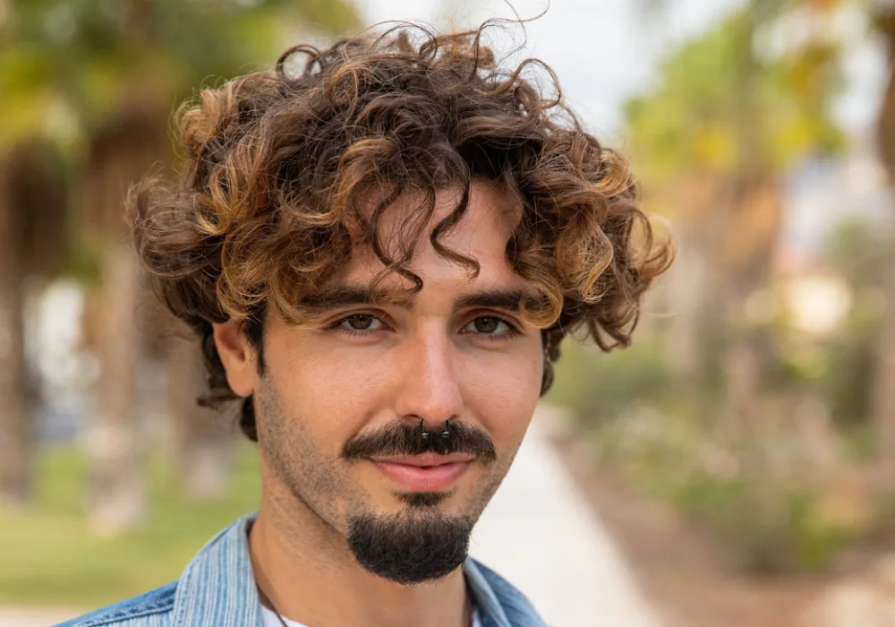 Smiling man with dark curly hair stands on a walking trail and wears a mustache and goatee combo to show one of the most popular goatee styles right now