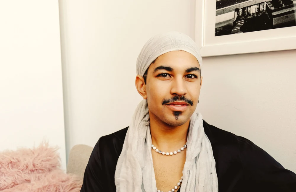 Nonbinary person sits smiling while wearing a long head scarf and necklace to show examples of goatee styles featuring a soul patch and mustache