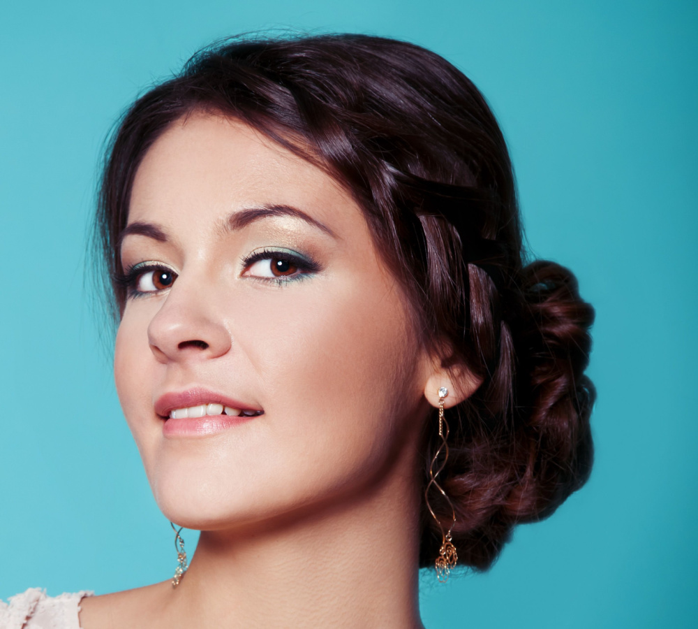 Brunette woman in front of a teal background smiles wearing a braided side bun as an example of trendy homecoming hairstyles