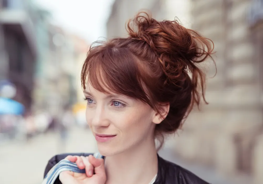 Red-haired woman with a high messy bun shows the concept of hair loss hairstyles for thinning hair on crown while walking in the city with a bag over her shoulder