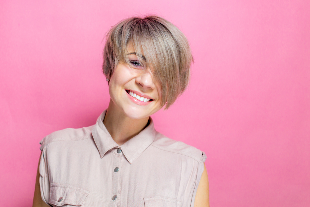 Woman smiles in front of a pink background with sleeveless top and razored pixie bob, an example of the best low maintenance haircuts for women