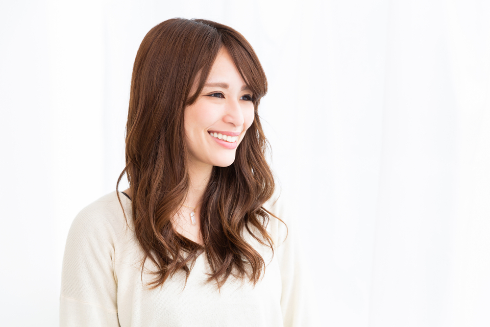 Asian woman smiles looking away with a white shirt and long layered hair with bangs, listed as one of our favorite low maintenance hairstyles for women