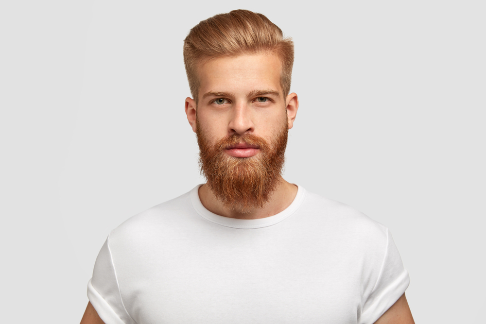 Man with dark blonde hair and beard wears a white t-shirt and smiles with a pompadour hairdo and chevron mustache with a full beard to show one of the most popular mustache styles