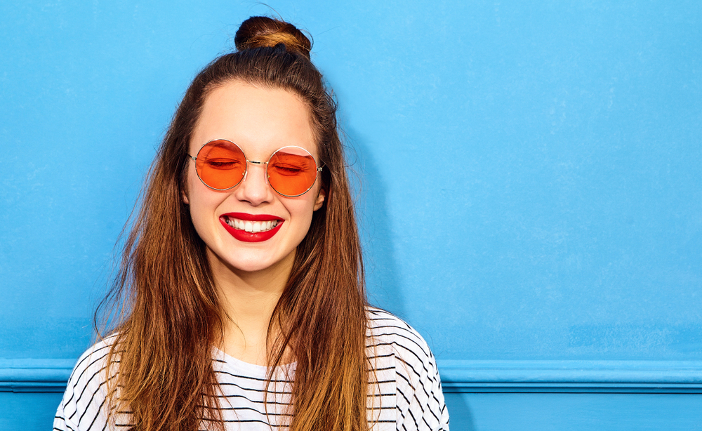 Young woman with orange sunglasses stands in front of a blue wall with a smile and closed eyes to show a great hair loss hairstyle for thinning hair on crown
