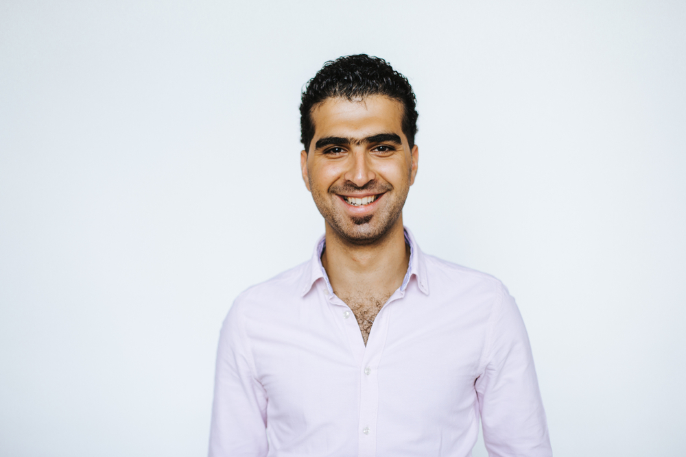 Arab man in a lilac dress shirt smiles brightly with a short haircut and soul patch goatee while standing in front of a white wall