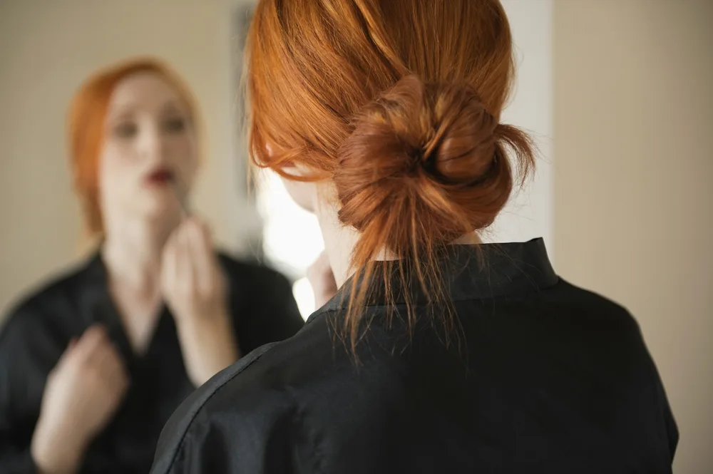 Back view of redhead woman wearing a low messy bun while looking in the mirror to show hairstyles for long straight hair