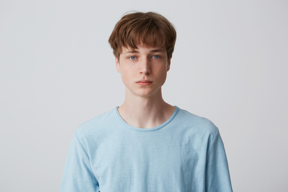 Young man in a blue shirt stares straight ahead in front of a white wall to model low maintenance haircuts for men with a choppy tapered cut