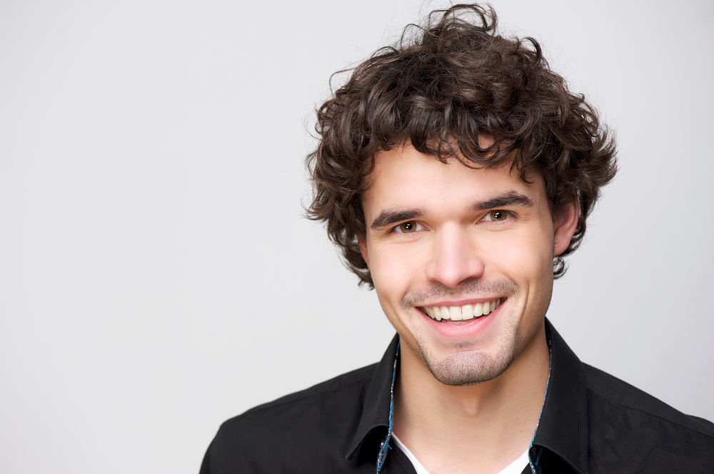 Curly-haired brunette man smiles with a curly mushroom cut that's been layered and tapered to be low maintenance