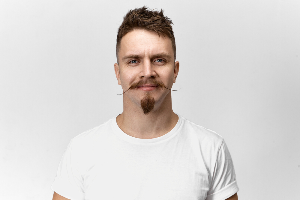 Man wearing a white t-shirt in front of a white wall shows off a Van Dyke goatee with handlebar mustache