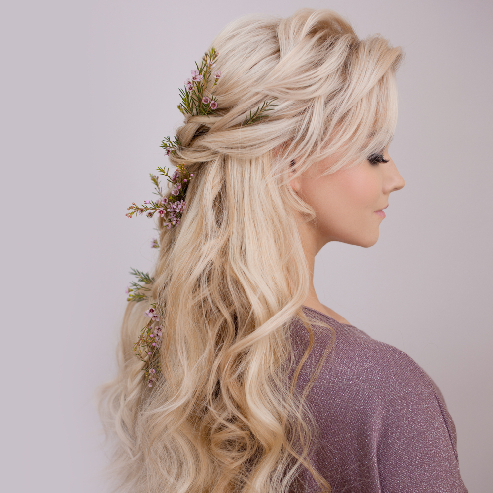 Profile view of young blonde woman modeling half-up homecoming hairstyles with curls and flower accents