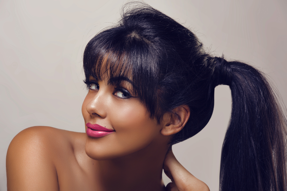 Black-haired woman leans over and smiles with bare shoulders to model one of the best ponytail hairstyles with hair wrapped around the base