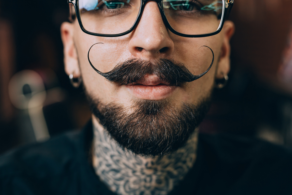 Close-up of hipster tattooed man with earrings wearing one of the 13 mustache styles, a handlebar mustache with waxed ends