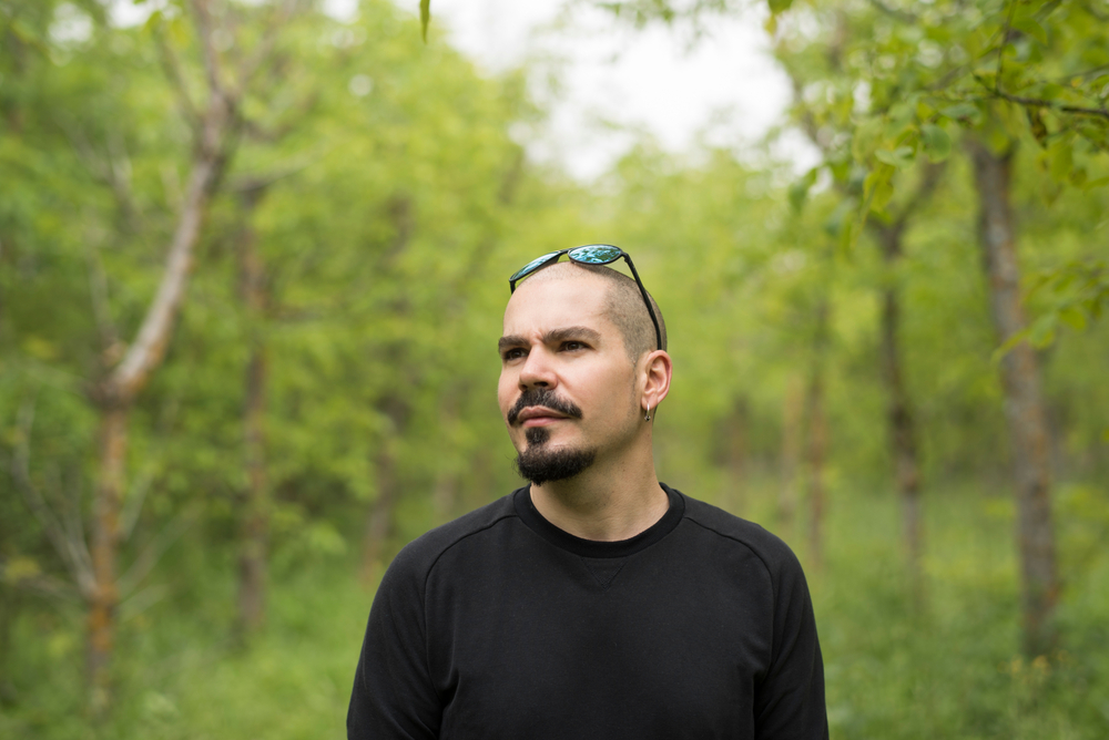 Man with buzzed haircut walks through the woods with sunglasses on his head and a black shirt, rocking a classic goatee with soul patch