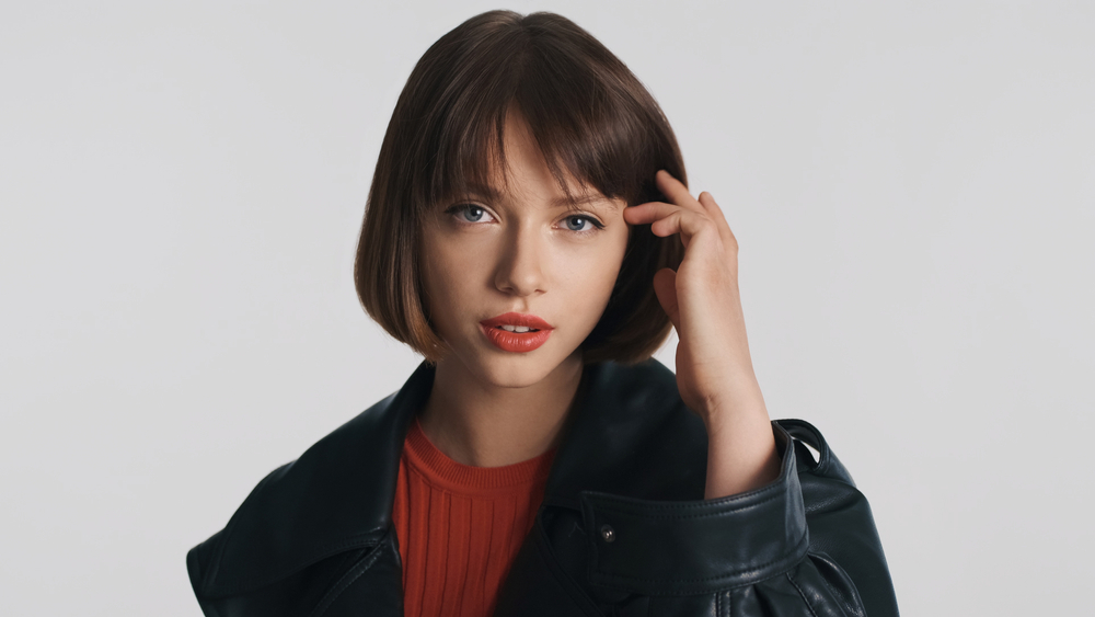 Brunette young woman with a French bob haircut for fine thin hair tucks her hair behind one ear with a leather jacket and red shirt on