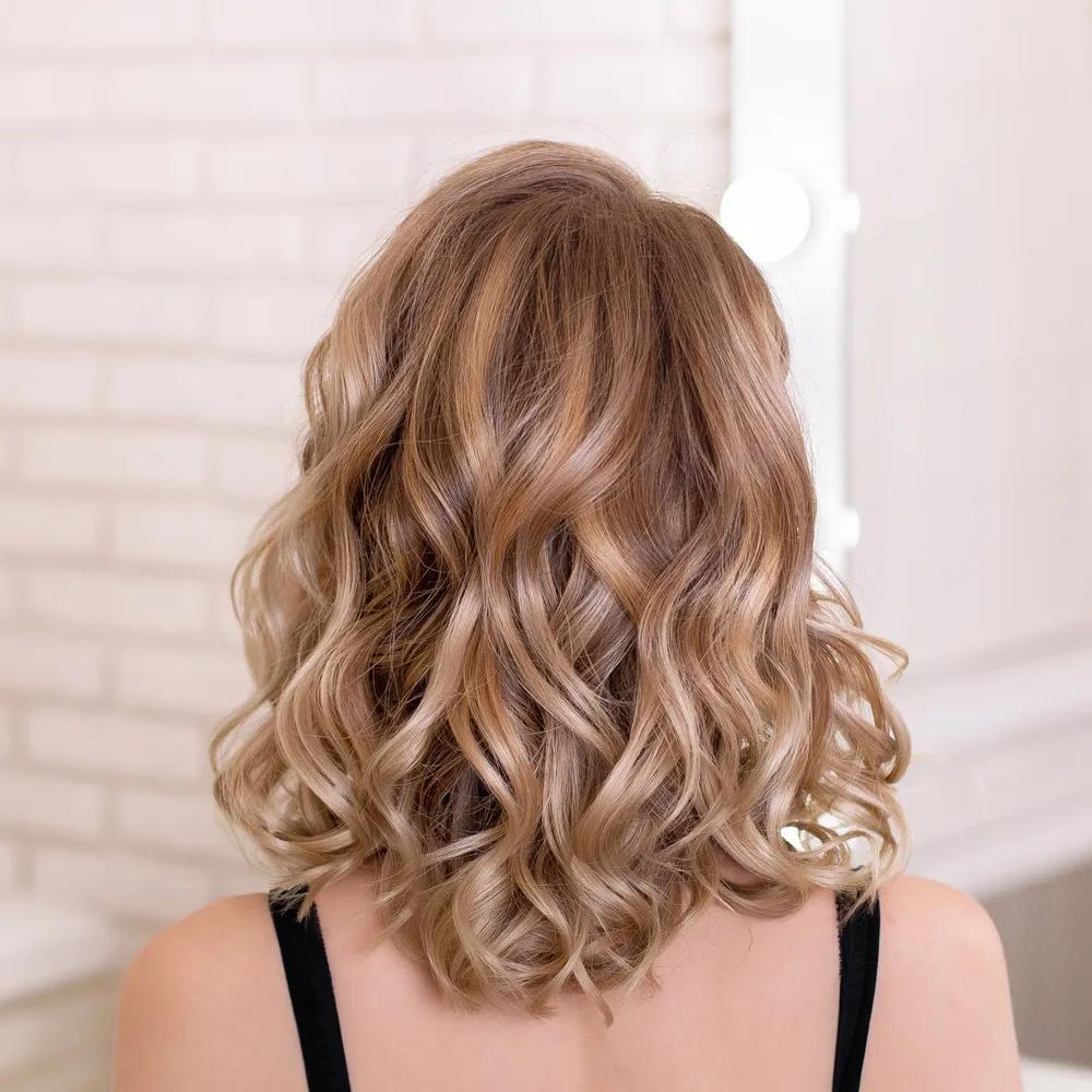 Rear view of a blonde woman with wavy hair standing in front of a white brick wall with a great hairstyle for thinning hair on the crown