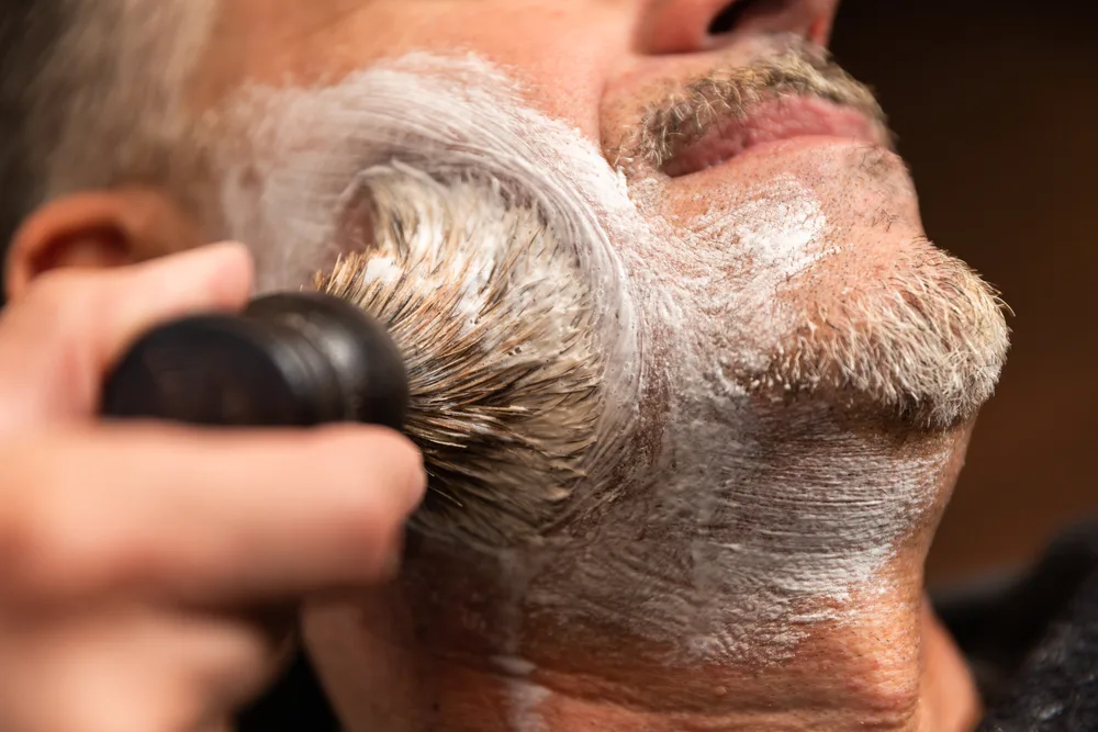Close-up image of barber applying shaving cream with a brush to a man's face with a goatee, showing the concept of trimming and grooming different goatee styles