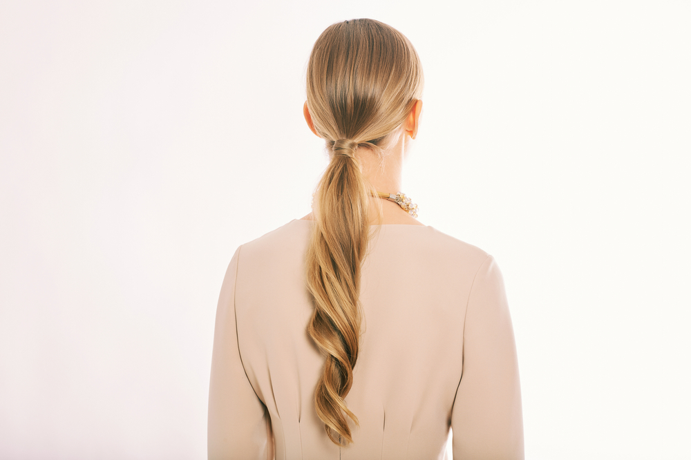 Back view of a blonde woman wearing a low ponytail with curled texture in front of a white wall