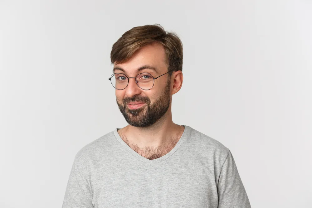Smiling man in glasses poses in front of a light gray wall with a tapered haircut that's longer on top and parted to the side