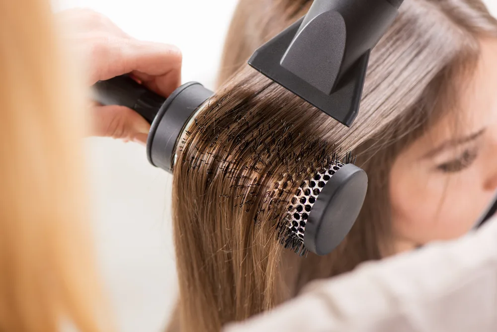 Woman with long hair gets a blowout with a round brush at a salon for a piece showing the best hairstyles for long straight hair