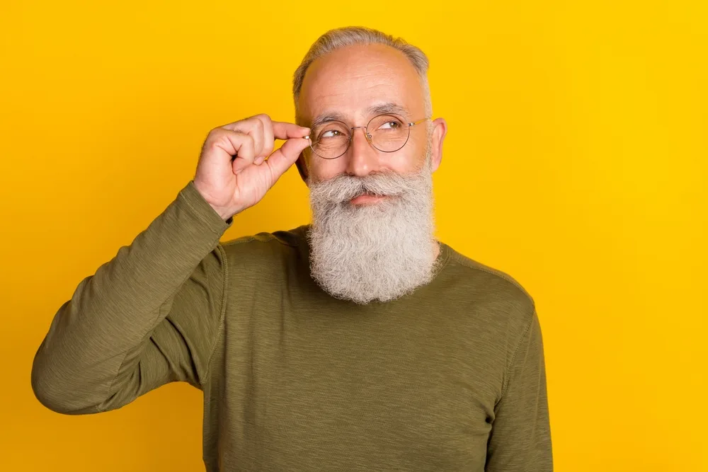 Gray-haired man in a green long-sleeved shirt touches his glasses with a Hungarian mustache and full beard in front of a yellow wall
