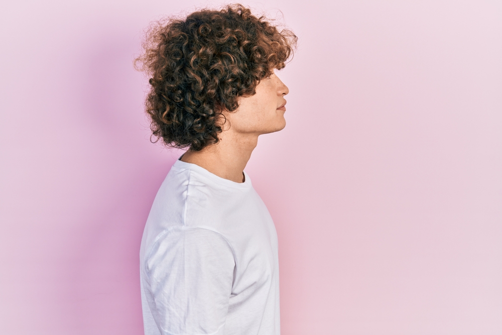 Young man with medium length curly hair has a low maintenance tapered haircut while standing in front of a pink wall with a long sleeved shirt
