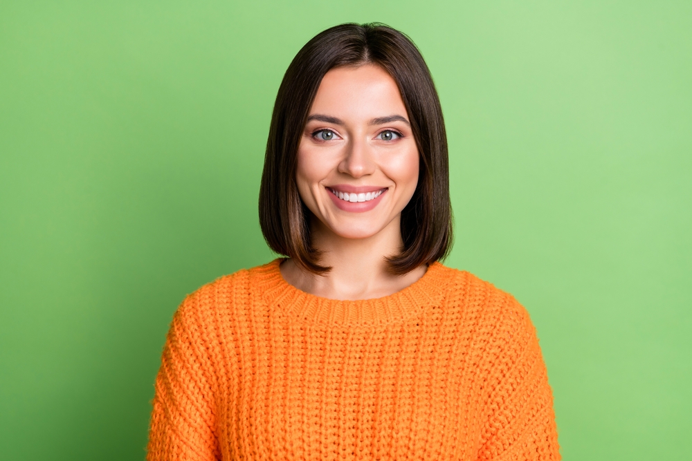 Brunette woman wearing an orange sweater smiles in front of a green wall with a long bob, representing the best low maintenance haircuts for women