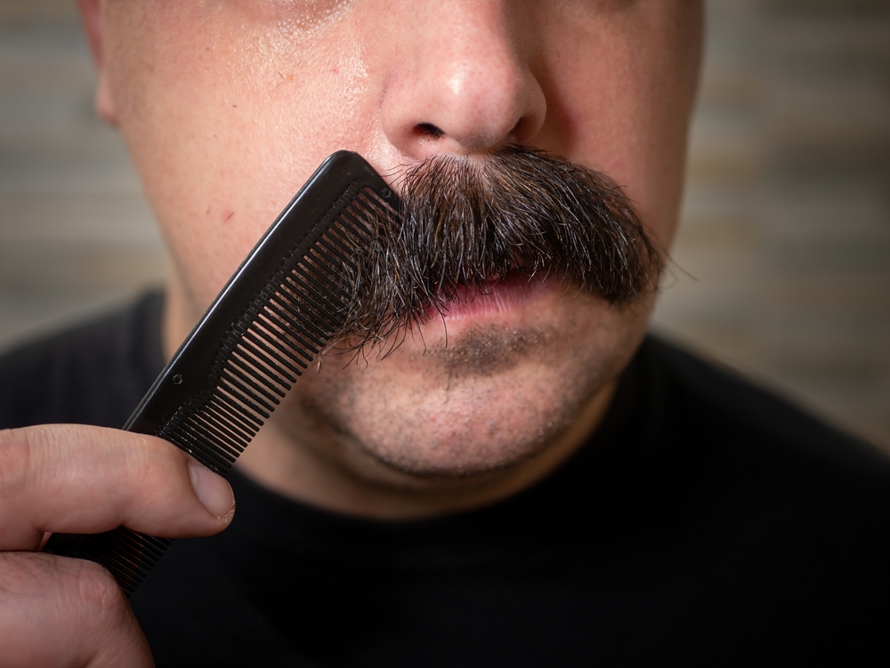 Close-up image of a man with a thick, brown walrus mustache combing the hair with a black t-shirt on
