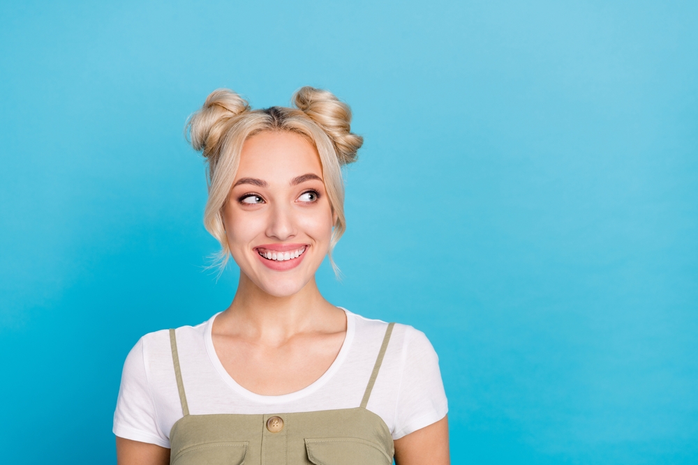 Blonde woman in front of a blue wall smiles with space buns hairstyle for long straight hair in a t-shirt and dress