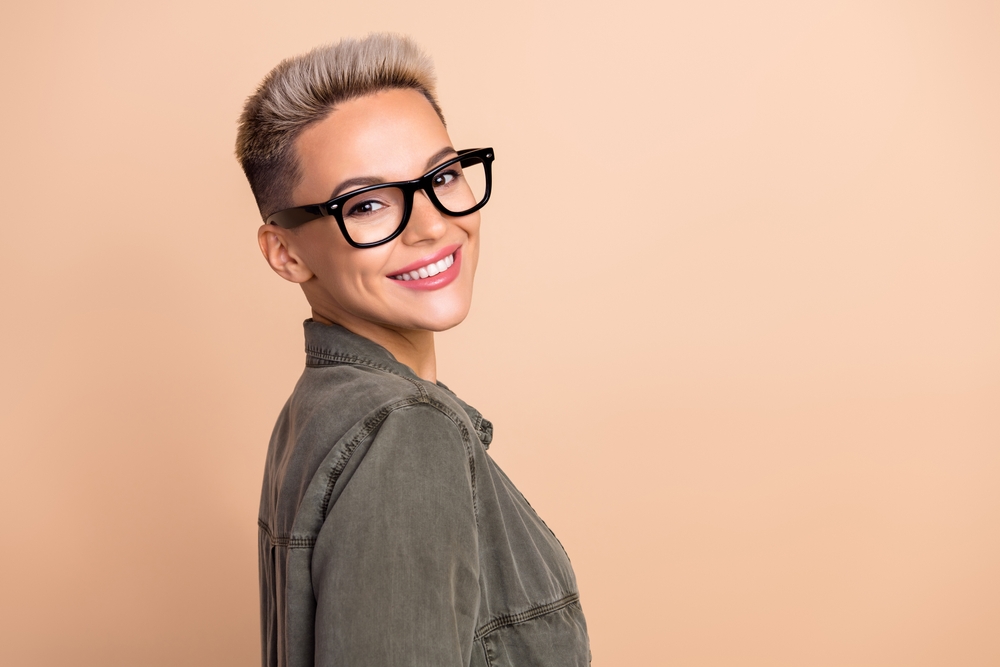 Woman in a green military style jacket smiles over her shoulder in front of a peach wall with glasses and a tapered pixie fade haircut for fine thin hair