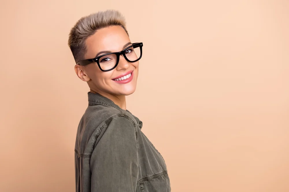 Woman in a green military style jacket smiles over her shoulder in front of a peach wall with glasses and a tapered pixie fade haircut for fine thin hair