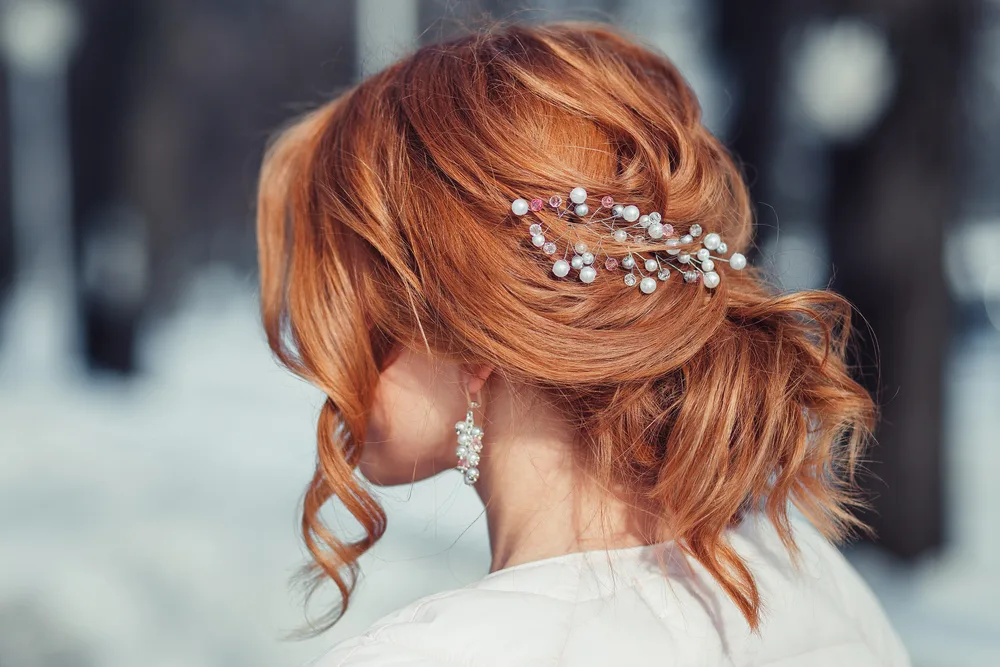 Back side view of red haired woman wearing a formal loose updo with pieces hanging loose and accessories pinned in the back